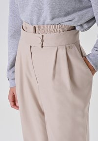 Pantaloni beige a vita alta con una larga cintura, dettaglio con bottoni, pieghe e tasche laterali, indossati con un top grigio chiaro a maniche lunghe.