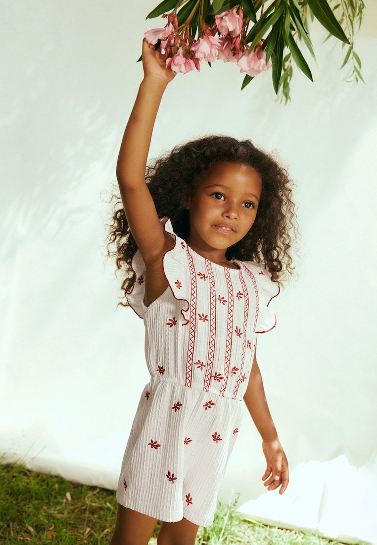 Fille portant une combinaison blanche avec une broderie rouge tend la main pour toucher des fleurs roses suspendues au-dessus de sa tête.