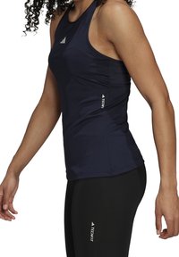 Žena nosící námořnickou modrou bezrukávovou sportovní top a černé legíny s logy "Techfit", zobrazená od ramen po boky.