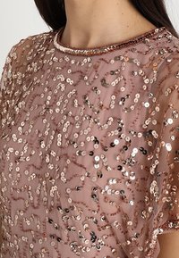 Przezroczysta bluzka w kolorze mauve z gęsto rozmieszczonymi cekinami w kolorze różowego złota i srebra w abstrakcyjnych, wirujących wzorach na krótkich rękawach.