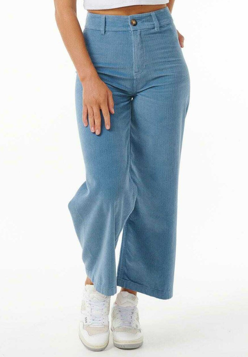 Helle blaue Corduroy-Hosen mit weitem Bein, hoher Taille, vorderem Knopfverschluss und vertikaler Rippenstruktur. Kombiniert mit weißen Sneakers.
