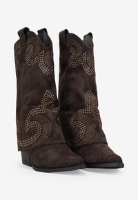 Botas de cowboy de ante marrón con recortes curvados y acentos de tachuelas metálicas en un patrón en espiral, con un tacón bajo y apilado y punta afilada.