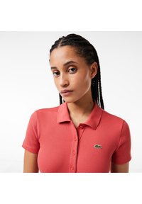 Camisa polo de canalé color coral con mangas cortas, cinco botones y un cuello redondeado. Presenta un pequeño logo verde de Lacoste en el lado izquierdo del pecho.