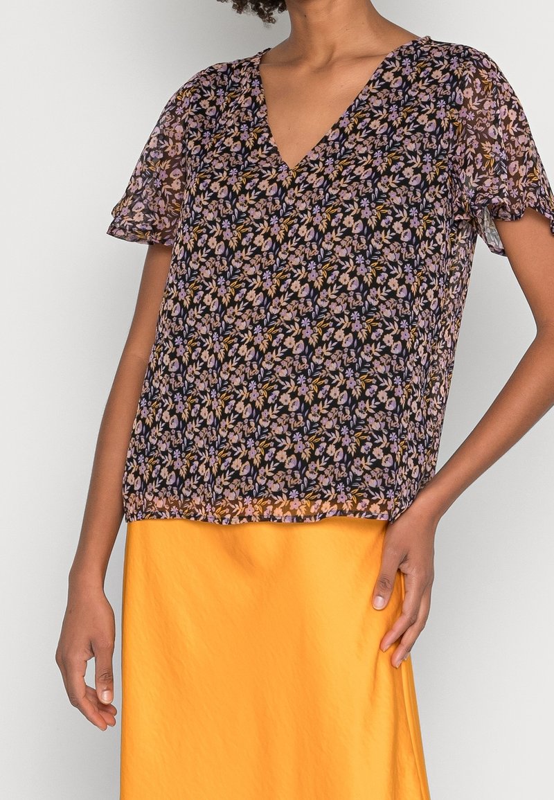 Femme portant un chemisier noir transparent à motifs floraux avec des manches courtes et une jupe jaune vif, debout, une main sur la hanche.