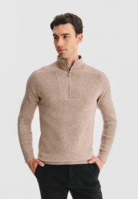 Beżowy sweter z mieszanki wełny z wysokim kołnierzem i półsuwnym zapięciem. Rękawy ze ściągaczami oraz teksturowana część korpusowa. Dopasowany krój.