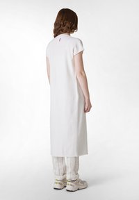 Weißes Leinenkleid mit kurzen Ärmeln, gerader Silhouette und einem kleinen lila Etikett auf der Rückseite, kombiniert mit gestreiften beigen Hosen und Sneakers.