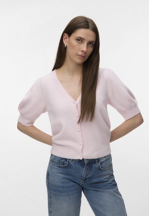 Vrouw met lang, stijl bruin haar, die een lichtroze vest met knopen en pofmouwen draagt, en een blauwe spijkerbroek, handen op de heupen.