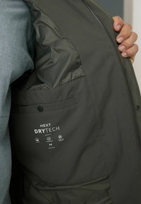 Mørkegrøn vest lavet af let, vandafvisende materiale. Har en lynlås, sidelomme og mærkeetiket "NEXT DRYTECH" indeni.