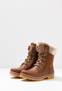 Botas de tobillo de cuero marrón y ante con forro de piel de oveja, diseño de cordones, ojales dorados y una suela de goma robusta. Logotipo en el costado.