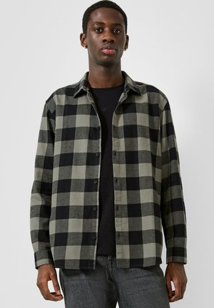 Mann trägt ein offenes schwarz-grau kariertes Langarmhemd über einem schwarzen T-Shirt und dunkelgraue Jeans, steht vor einem einfarbigen Hintergrund.