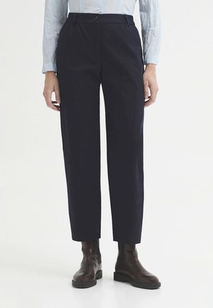 Personne portant un pantalon droit bleu marine foncé, une chemise à manches longues bleu clair rentrée, et des bottines marron foncé sur fond blanc.