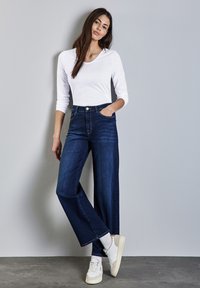 Dunkelblaue weite Jeans mit Fronttaschen, kombiniert mit einem weißen Langarmoberteil und beigefarbenen Sneakers vor einem grauen Hintergrund.