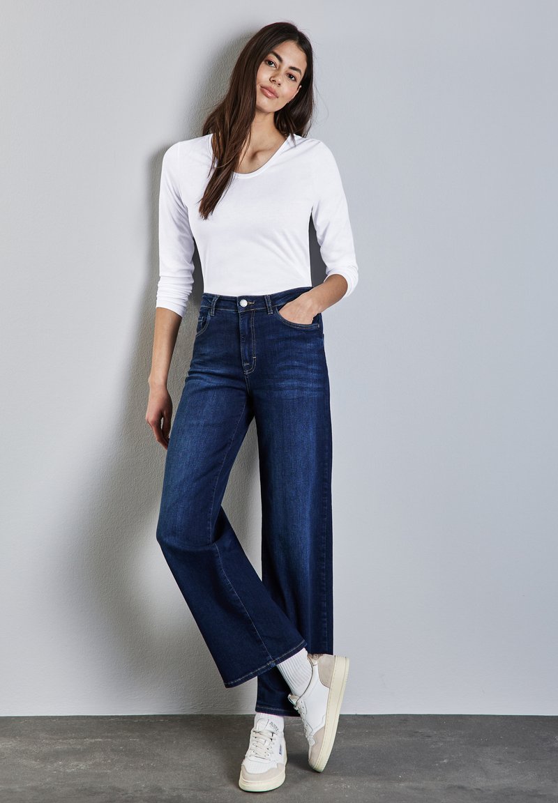 Dunkelblaue weite Jeans mit Fronttaschen, kombiniert mit einem weißen Langarmoberteil und beigefarbenen Sneakers vor einem grauen Hintergrund.