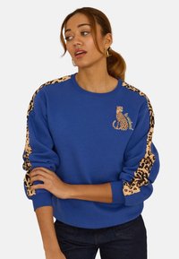 Blauwe trui met luipaardprintaccenten op de mouwen en een geborduurd luipaardontwerp op de borst. Zachte stof en een ontspannen pasvorm.