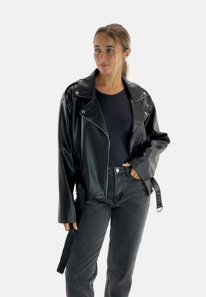 Veste de biker en simili cuir noir avec des garnitures argentées, grands revers et une ceinture ajustable. Portée par-dessus un haut noir et un jean foncé.