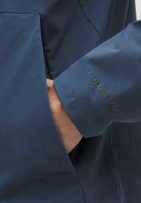 Hand in een donkerblauwe Lululemon-jack met de hand in de voorzak van de jas.