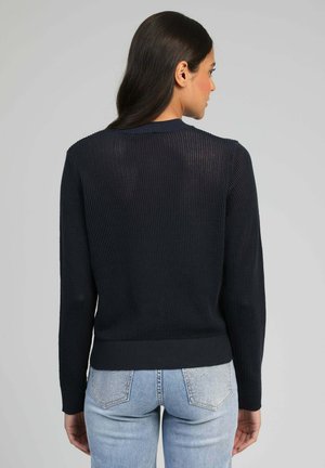 Femme aux longs cheveux bruns portant un pull en tricot noir et un jean bleu clair, debout de dos contre un fond gris uni.