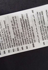 Etichetta di cura tessuta, tessuto bianco con testo nero in più lingue. Include la composizione del materiale: 70% cotone, 30% poliestere riciclato.