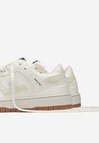 White sneakers made of leather with a textured finish, featuring a gum rubber outsole, flat laces, and gold branding on the side.