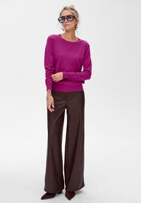 Kobieta w dużych ciemnych okularach przeciwsłonecznych, magentowym swetrze, szerokich brązowych skórzanych spodniach i brązowych czółenkach, stojąca na białym tle.