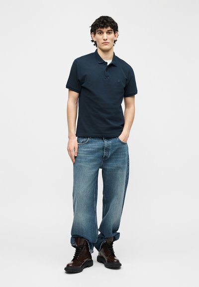 Νεαρός άνδρας που στέκεται με τα χέρια στις τσέπες, φοράει μπλε πόλο πουκάμισο, φαρδιά μπλε τζιν με γυρισμένα μπατζάκια στους αστραγάλους και σκούρα καφέ μπότες με κορδόνια, σε λευκό φόντο.