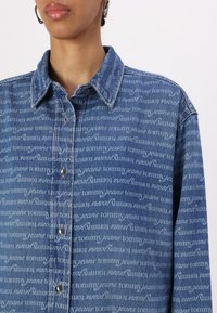 Sininen farkkupaita, jossa valkoinen "tommy jeans" -tekstikuvio koko pinnalla, klassinen kaulus, napillinen edessä ja pitkät hihat. Karkea kangas.