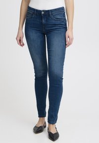 Jeans ajustados en denim azul oscuro, con cintura de tiro medio, desvanecido mínimo y cierre clásico con botón. Combinados con zapatos planos negros.