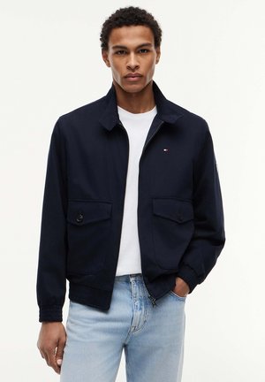 Giovane uomo che indossa una giacca blu navy con cerniera sopra una maglietta bianca e jeans azzurri, in piedi con una mano in tasca su sfondo bianco.