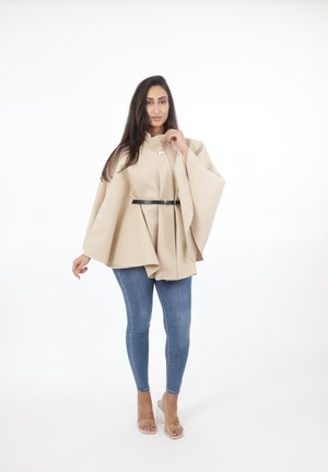 Beige struktureret kappe med klokkeærmer, fastgjort med et sort bælte om taljen. Sammen med lyseblå tætsiddende jeans og gennemsigtige hæle.