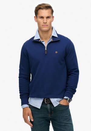 Navy pullover met een kwart rits en klein logo, gelaagd over een lichtblauw gestreefd shirt. Heeft ribgebreide manchetten en een ontspannen pasvorm.