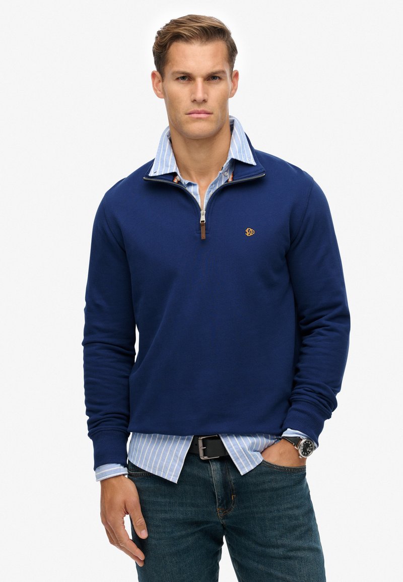 Jersey azul marino con cremallera de cuarto y un pequeño logo, sobre una camisa a rayas azul claro. Presenta puños acanalados y un corte relajado.