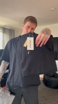 Sudadera negra hecha de tela suave, con un cuello redondo y etiquetas adjuntas. Presentada con una camiseta gris en la otra mano.