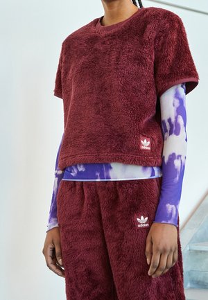 Personne portant un haut à manches courtes en peluche bordeaux de la marque Adidas et un pantalon, par-dessus un t-shirt à manches longues tie-dye violet et blanc, debout à l'intérieur.