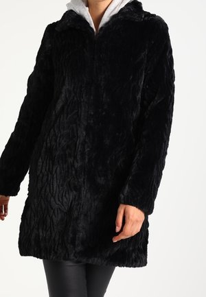 Femme portant un manteau en fausse fourrure noire texturée par-dessus un sweat à capuche gris clair et un pantalon noir.