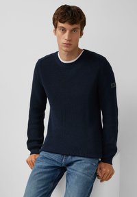 Dunkelblauer gerippter Strickpullover mit Rundhalsausschnitt und langen Ärmeln, der eine strukturierte Oberfläche und ein Logopatch am linken Arm aufweist.