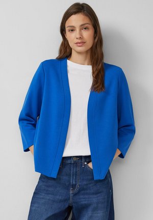 Kobieta o długich brązowych włosach, ubrana w jasnoniebieski kardigan z otwartym przodem, białą koszulkę i niebieskie jeansy, stojąca z rękami w kieszeniach.