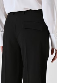 Pantaloni neri sartoriali in tessuto liscio, con due tasche posteriori e una vita su misura, indossati con una maglietta bianca a maniche lunghe.
