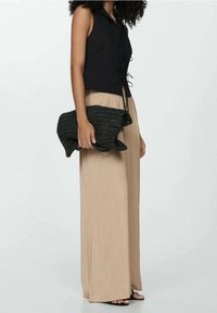 Blouse noire sans manches avec liens à l'avant, associée à un pantalon beige plissé, long jusqu'au sol. Tenant une pochette noire tissée texturée.