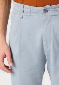 Pantalons bleu clair avec un motif en chevrons, dotés d'une fermeture à bouton et de passants de ceinture. Le tissu a une texture lisse et subtile.
