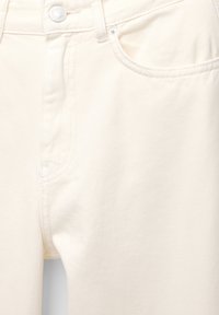 Jeans couleur crème avec une texture lisse, dotés d'une poche avant, d'une fermeture à bouton en métal et de détails de couture subtils.