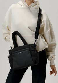 Svart vadderad axelväska med dubbla bärhandtag och justerbar rem, med en mjuk textur och logodetalj på framsidan.