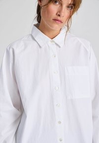 Camicia bianca a maniche lunghe in cotone, con chiusura a bottoni sul davanti, colletto a punta e una tasca sul petto. Texture liscia, vestibilità rilassata.
