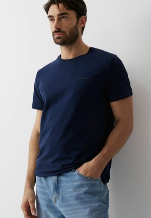 Mężczyzna o ciemnych włosach i brodzie, ubrany w granatowy t-shirt z krótkim rękawem i jasnoniebieskie dżinsy, stoi przy białej ścianie z jedną ręką w kieszeni.