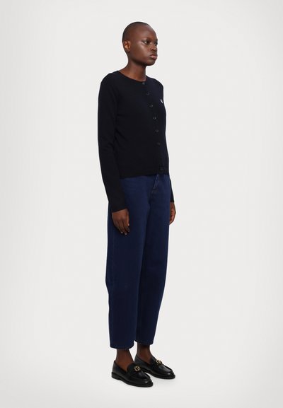 Svart knapp-up cardigan med lange ermer, laget av mykt materiale, kombinert med løse marineblå bukser og svarte loafers med gullakenter.
