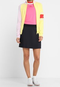 Sportovní outfit s žlutou a růžovou bundou na zip, růžovým tričkem a černou plisovanou sukní. Celý vzhled doplňují bílé sportovní boty.