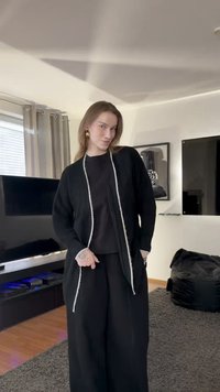 Czarny kardigan dziany z białym obszyciem, luźny krój, z odkrytym przodem, noszony na czarnym topie i luźnych czarnych spodniach, na tle neutralnego wnętrza.