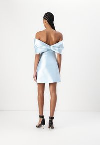 Mini abito in satin di colore azzurro chiaro con dettaglio a fiocco spalline scoperte sul retro. Presenta una silhouette aderente e una texture liscia. Sandali neri con tacco.