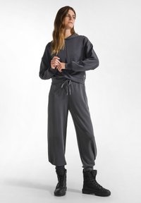 Grau Hoodie mit Kordelzug, kombiniert mit passenden lockeren Jogginghosen. Das Model trägt schwarze Schnürstiefel bis zu den Knöcheln. Glatte Stoffoberfläche.