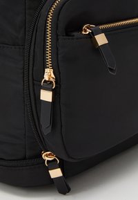 Svart tygväska med tre synliga gulddragkedjor och dragkedjehandtag i svart läder med guldaccenter.