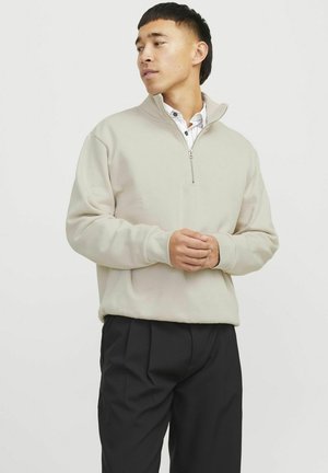 Pullover in pile beige chiaro con zip parziale e maniche lunghe, polsini a coste e una texture liscia, abbinato a pantaloni neri.